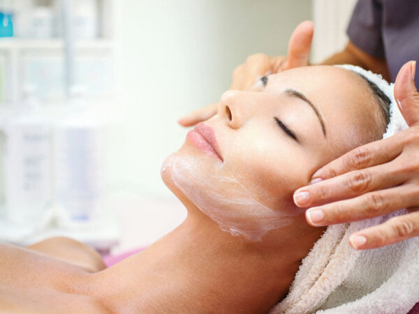 Closeup of adult caucasian woman having her face pampered at beauty salon by unrecognizable mid aged professional female beautician. The client is relaxing on massage bed wrapped in white towel. Some cosmetic products are on the shelves in background, blurry.